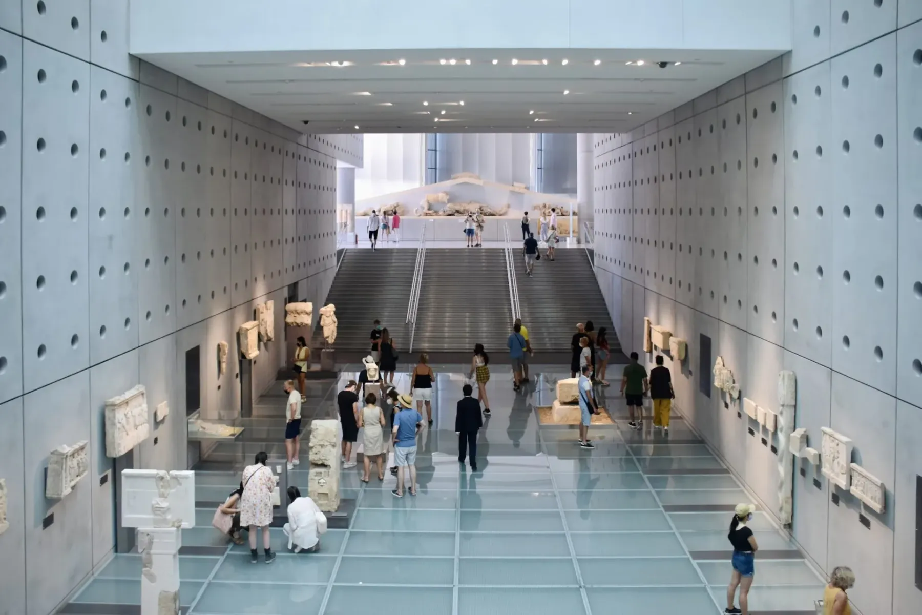 Innenansicht eines modernen Museums mit antiken Skulpturen an Betonwänden, Glasboden und Besuchern. Eine große Treppe führ...