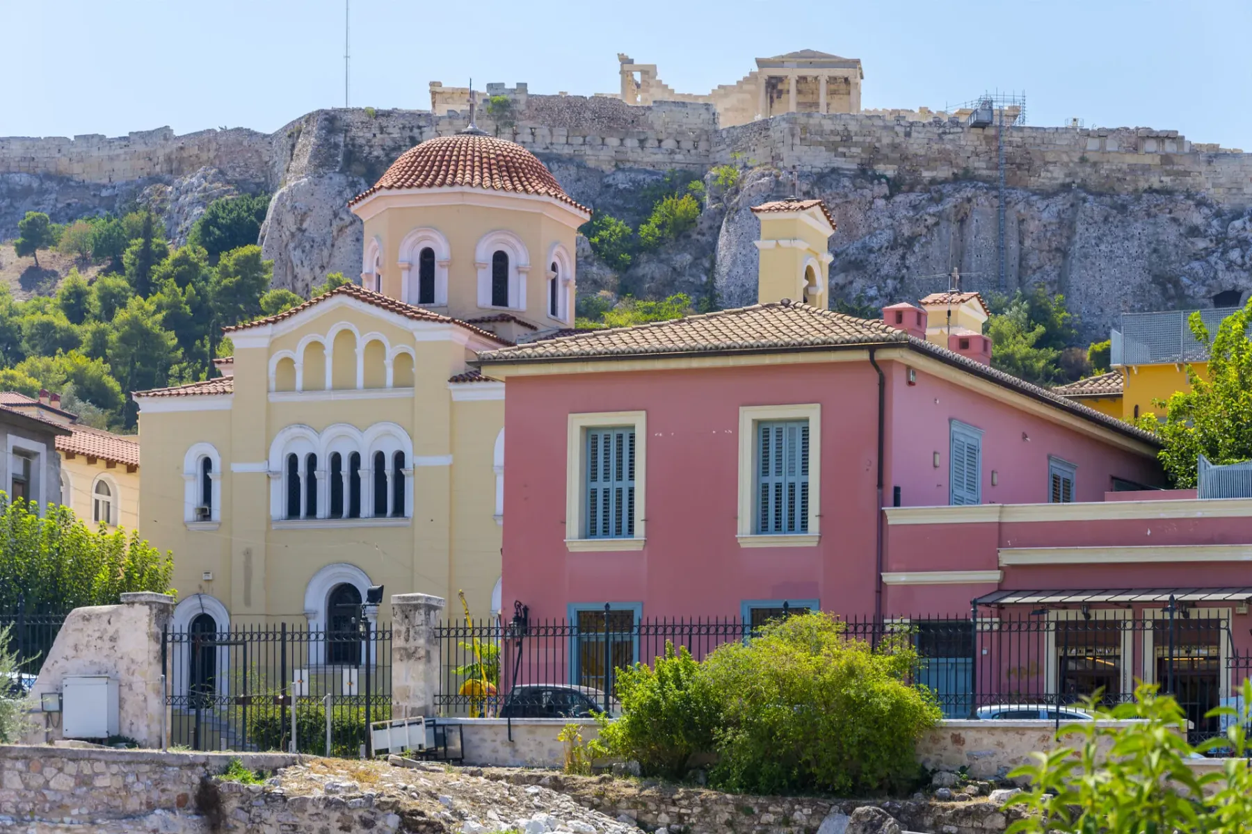 Gelbe Kuppelkirche und ein rosafarbenes Gebäude in Athen, mit dem felsigen Akropolis-Hügel und Parthenon-Ruinen im Hinterg...