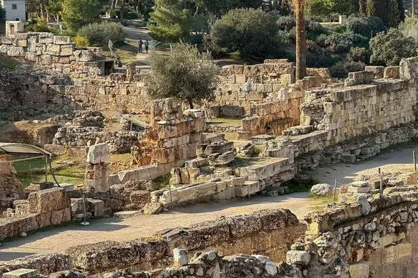 Extensive ancient stone ruins and walls with overgrown sections and dirt paths, surrounded by trees under a clear sky.