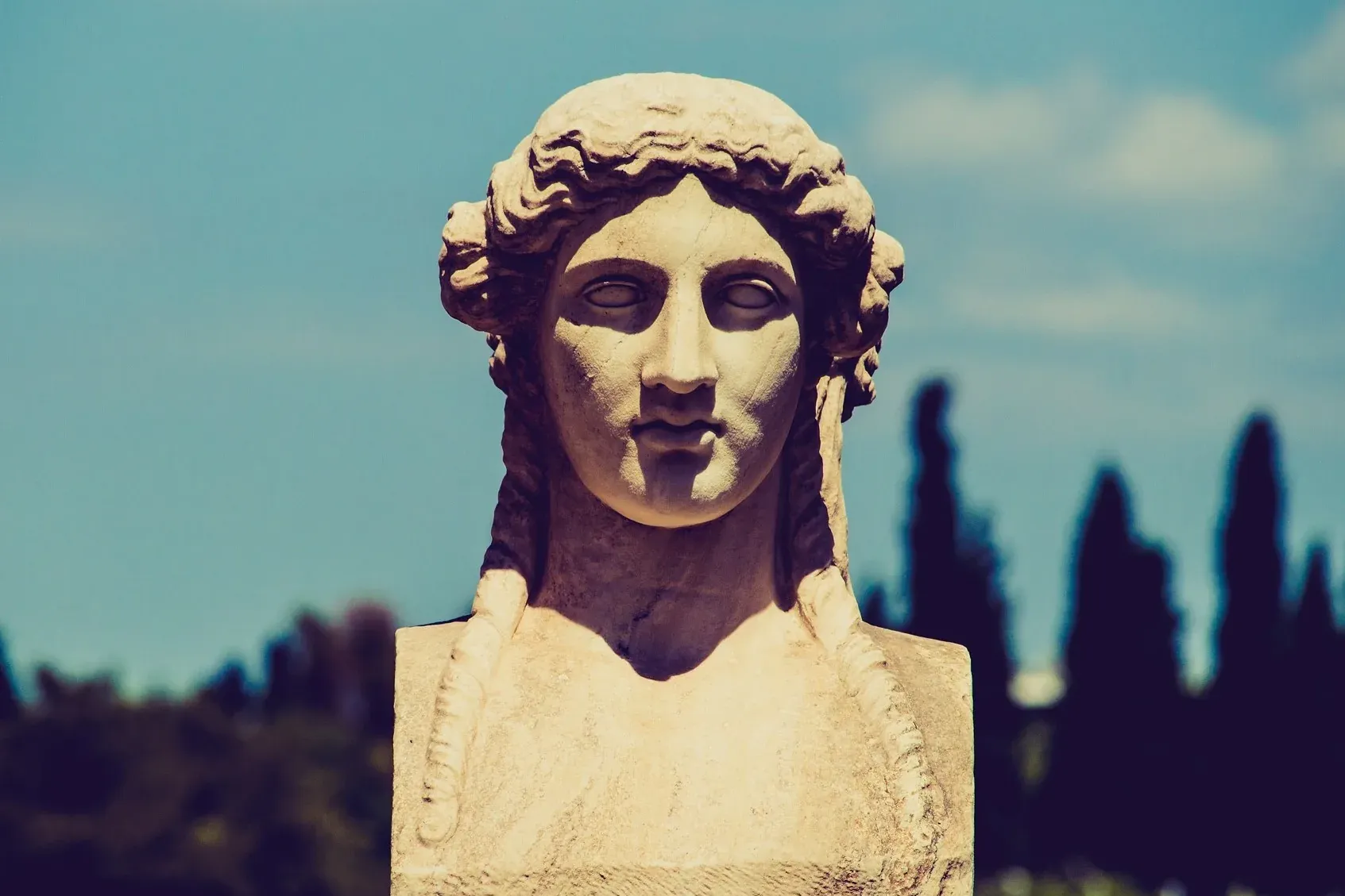 Nahaufnahme einer antiken griechischen Marmorbüste einer Frau mit geflochtenem Haar, vor klarem blauem Himmel.