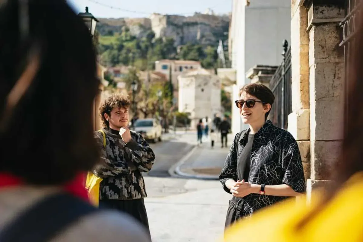 Lächelnde Reiseleiterin mit Sonnenbrille spricht mit einer kleinen Touristengruppe auf sonniger Straße in Athen, Akropolis...