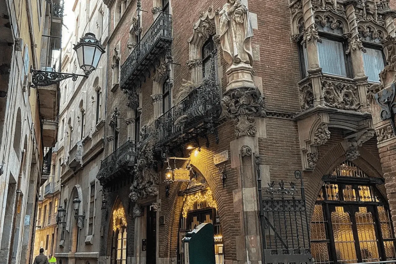 Edificio de ladrillo ornamentado con detalles de piedra tallada, estatua de una figura y balcones en una calle estrecha de...