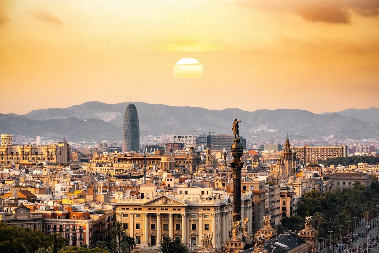 Vista panorámica de Barcelona al atardecer con el Monumento a Colón en primer plano y la Torre Glòries contra montañas.