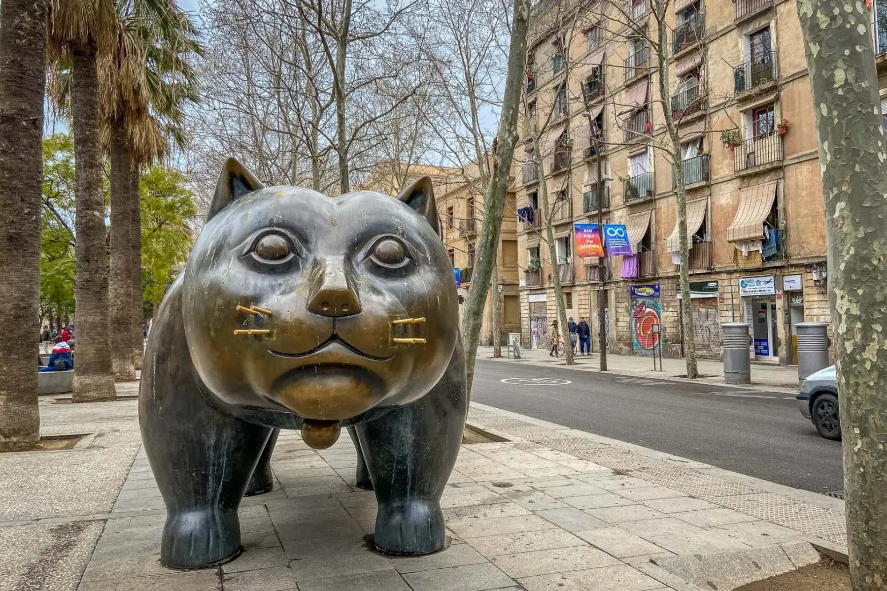 Große Bronzeskulptur einer pummeligen Katze auf einem Bürgersteig neben einer Straße mit Gebäuden und Bäumen.