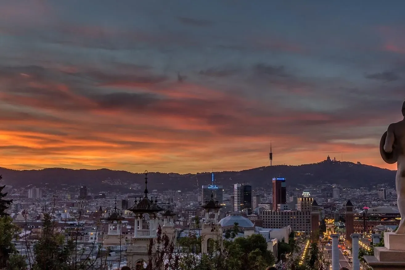 Vista del horizonte de Barcelona al atardecer con una estatua en primer plano y la montaña Tibidabo al fondo.