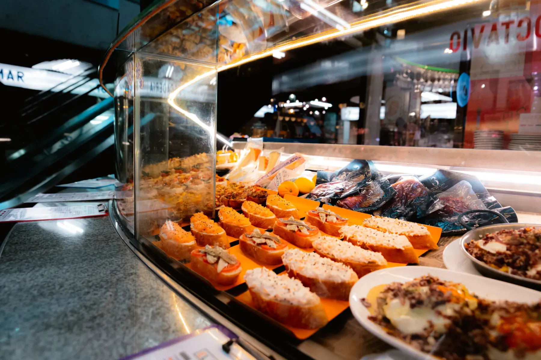 Food display counter with various pintxos, tapas, and packaged items under warm lighting.