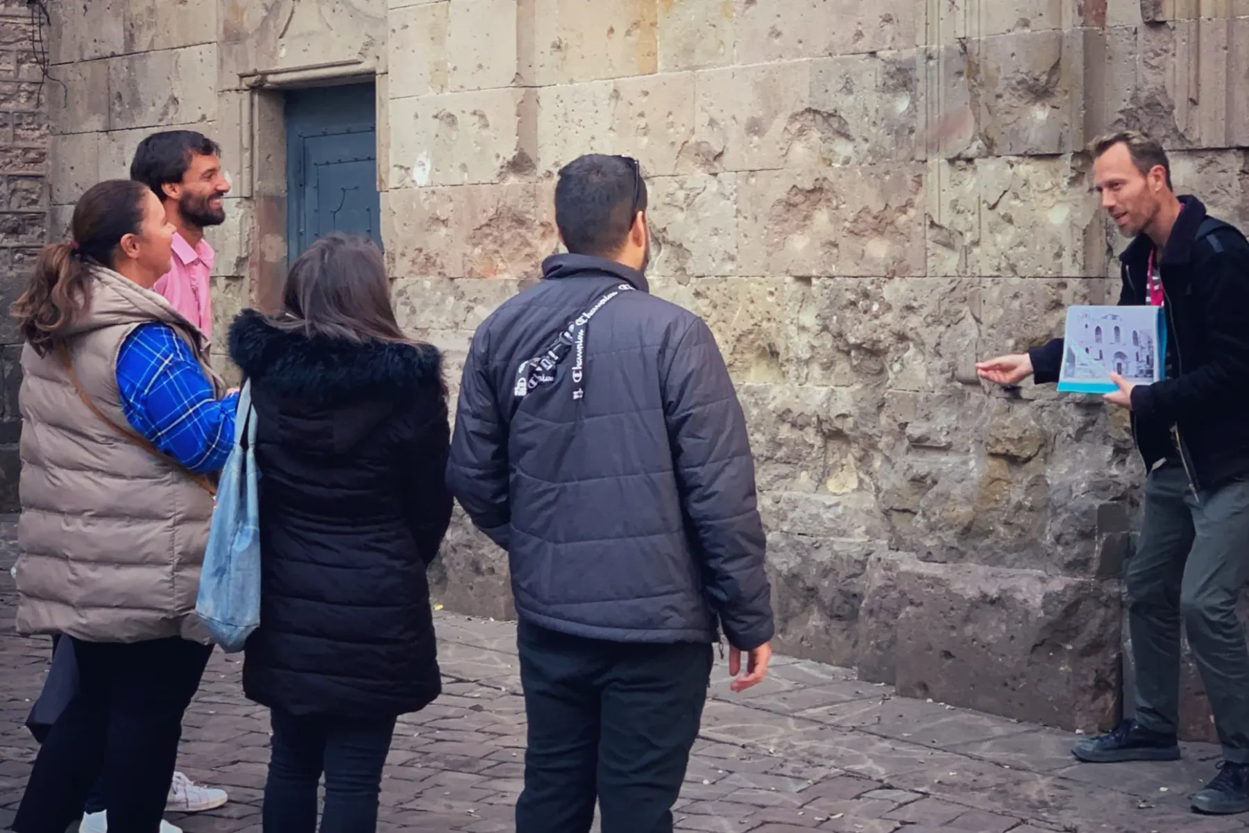 Ein männlicher Reiseführer mit Broschüre zeigt auf eine alte Steinmauer und spricht mit drei Touristen.