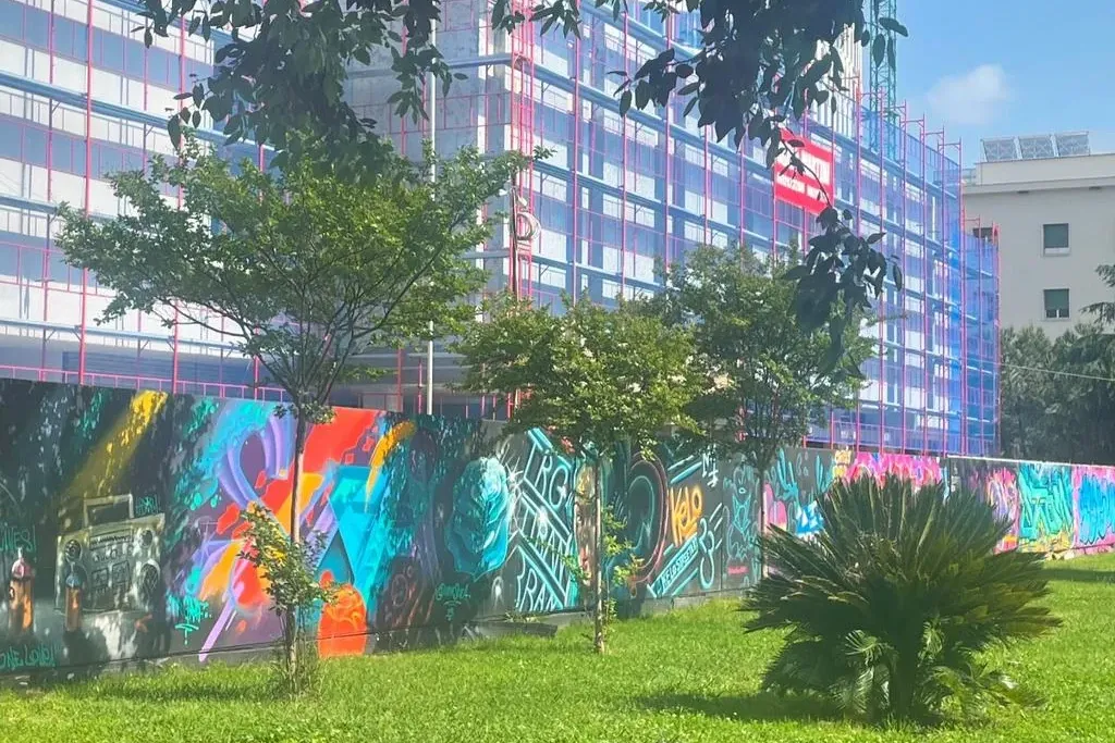 Kolorowy mural street artowy na murze z zieloną trawą na pierwszym planie, budynkiem w budowie i drzewami.