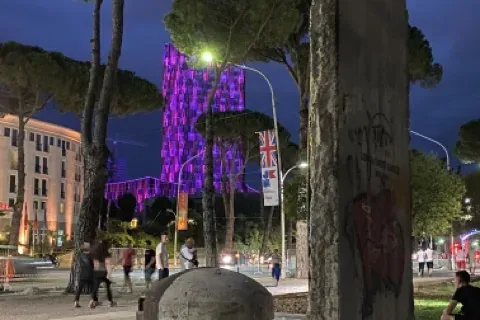 Betonowy bunkier i fragment Muru Berlińskiego z graffiti na placu w Tiranie o zmierzchu. W tle przechodzą ludzie.