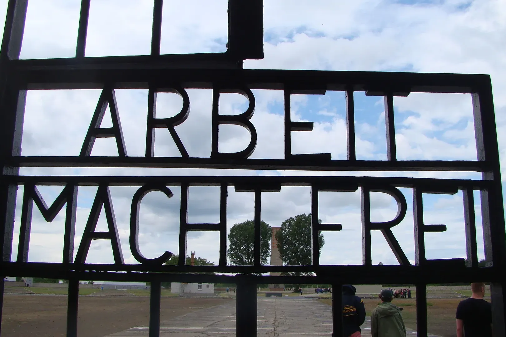 Dunkles Metalltor mit dem deutschen Schriftzug 'ARBEIT MACHT FREI' vor bewölktem Himmel an der Gedenkstätte Sachsenhausen.