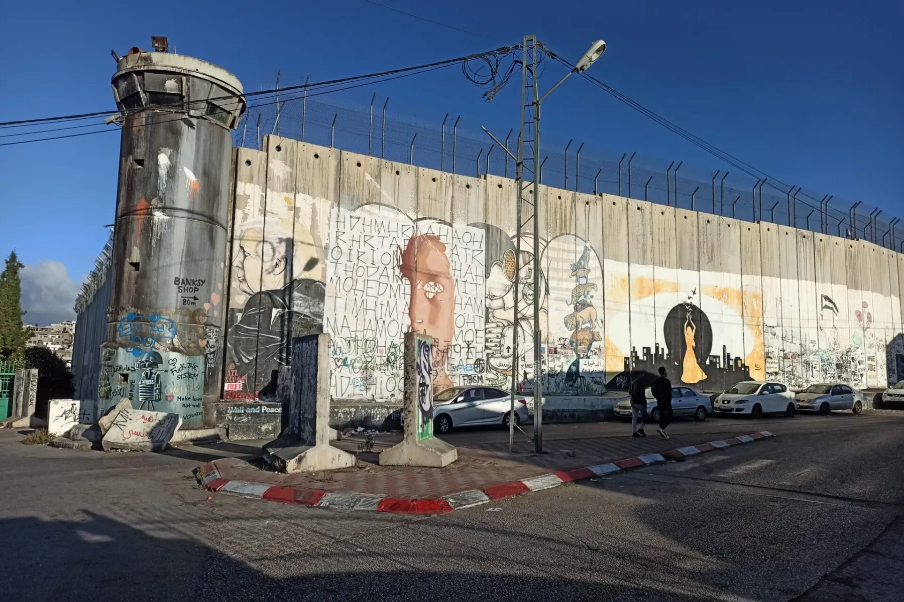 Muro de separación de hormigón con torre de vigilancia cubierto de grafitis y murales, coches aparcados, bajo cielo azul e...