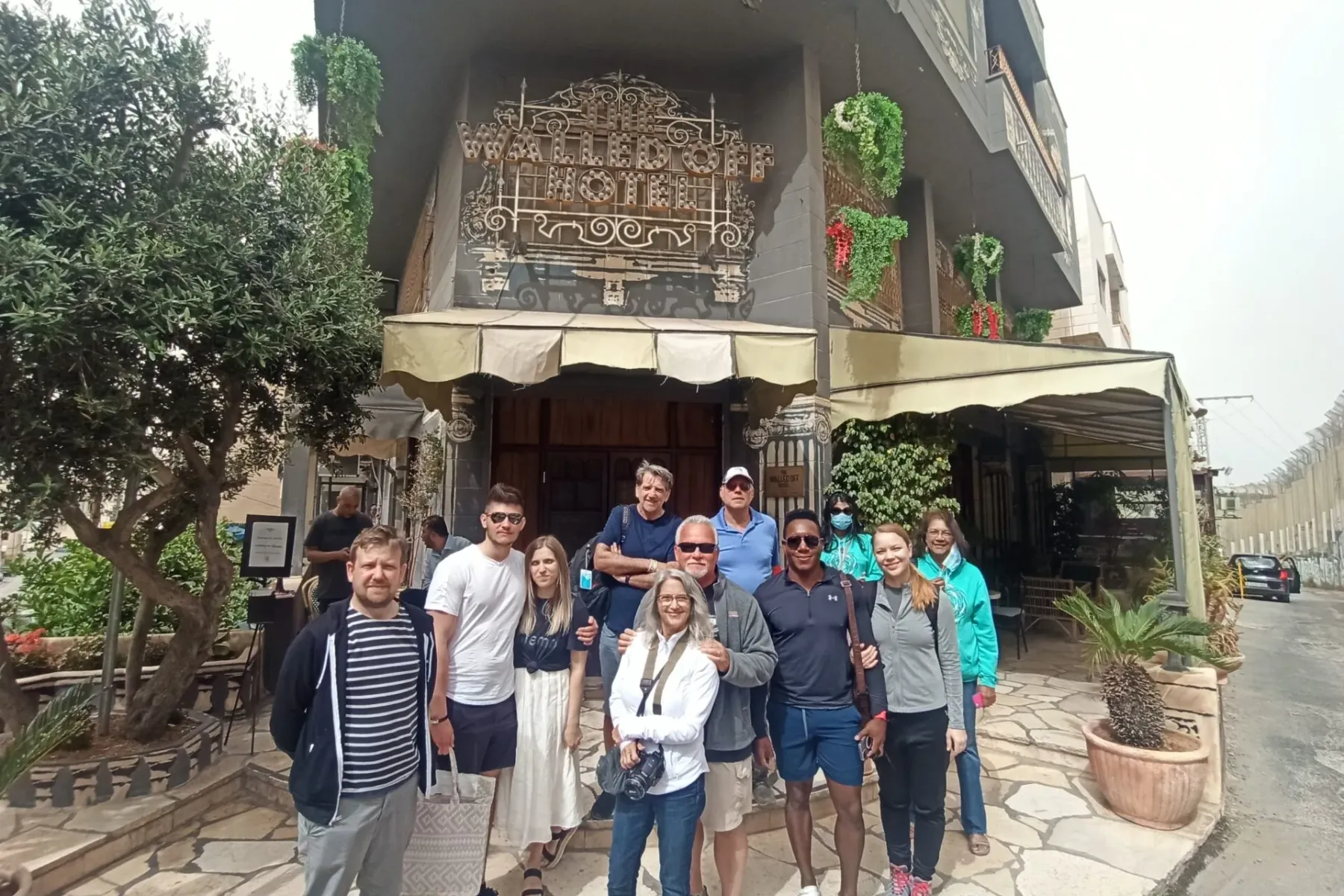 Grupo de diez turistas posando frente al The Walled Off Hotel en Belén, con un olivo y plantas en macetas.