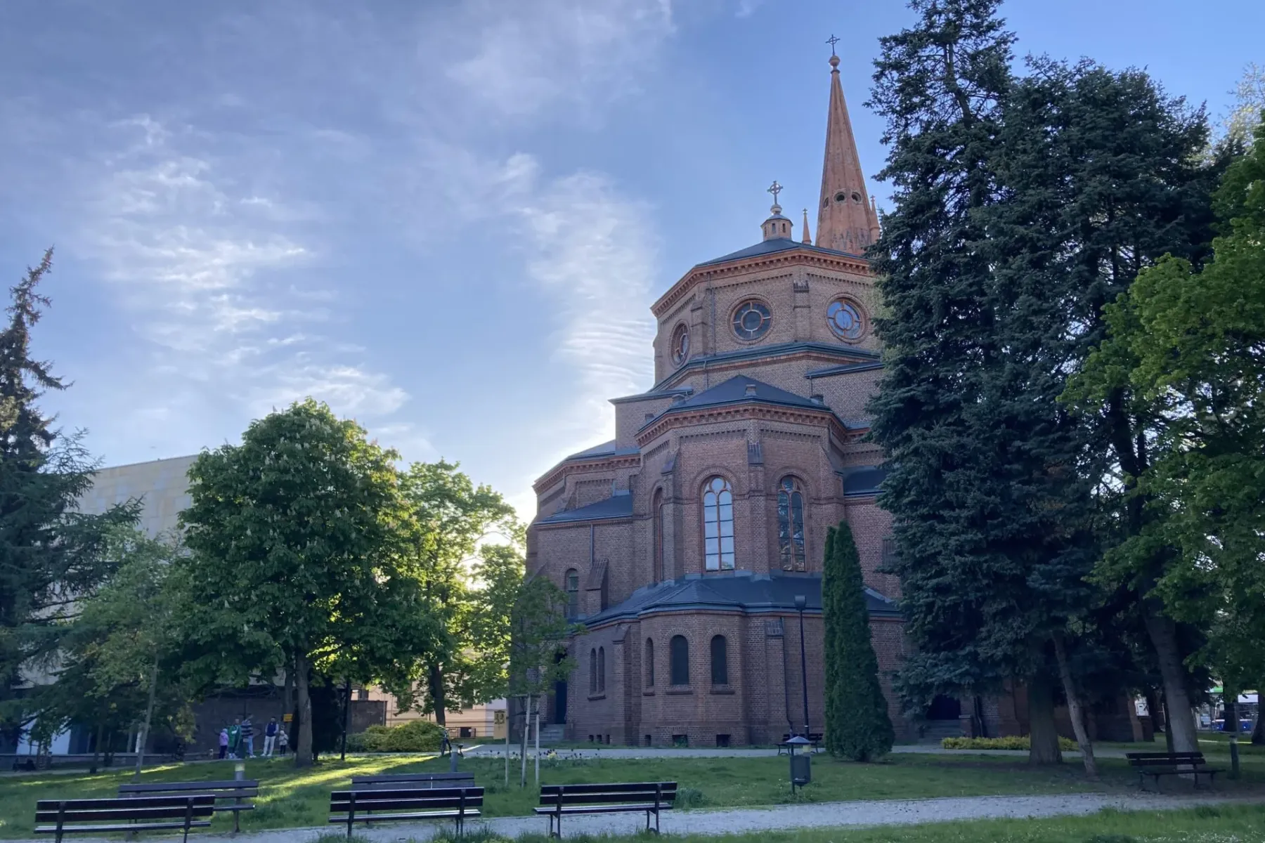 Große Backsteinkirche mit hohem Turm, von einem grünen Park mit Bäumen und Bänken unter teils bewölktem Himmel gesehen.