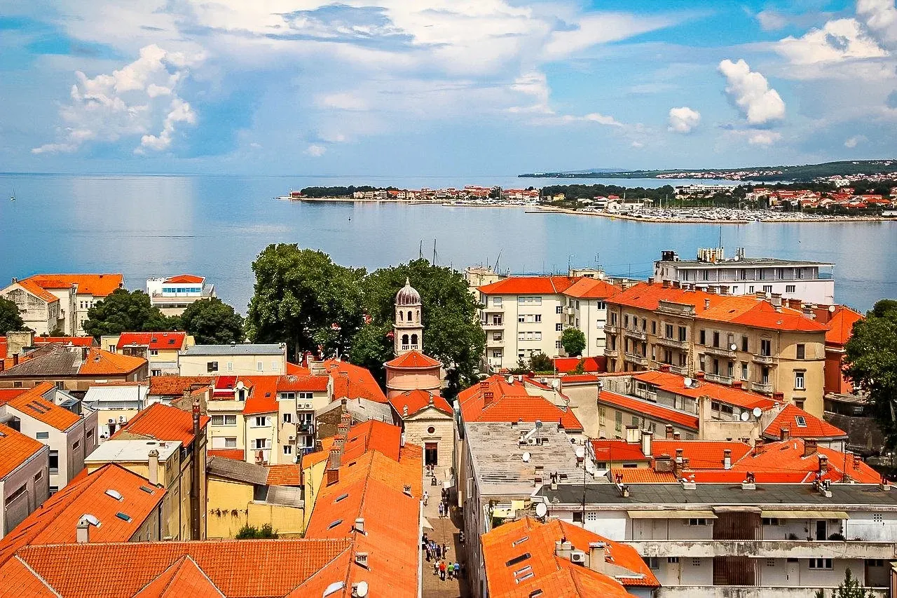 Vista aérea del casco antiguo de Zadar con tejados de tejas naranjas, un campanario abovedado y el mar Adriático.