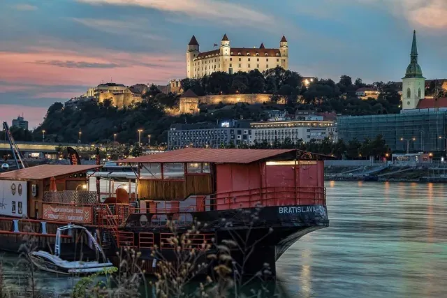 Castillo de Bratislava y Catedral de San Martín iluminados al atardecer. Bar flotante en primer plano sobre el Danubio.