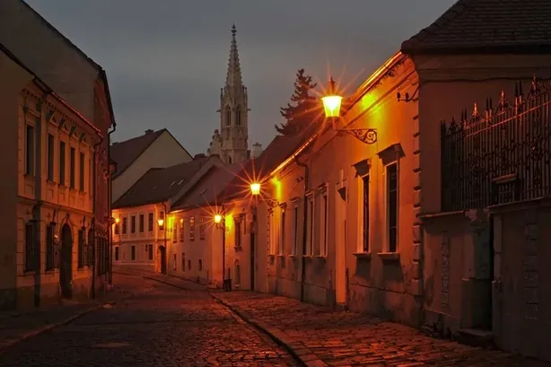 Calle empedrada con edificios antiguos de noche, iluminada por farolas cálidas, con un campanario de iglesia al fondo.