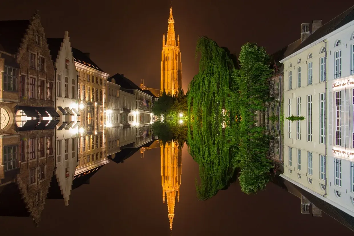 Oświetlone zabytkowe budynki i wieża Belfry odbijające się w spokojnym kanale w Brugii nocą.
