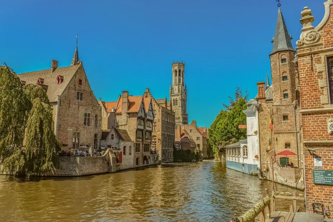A canal in Bruges with traditional brick buildings on both sides under a clear blue sky. The Belfry of Bruges is visible i...