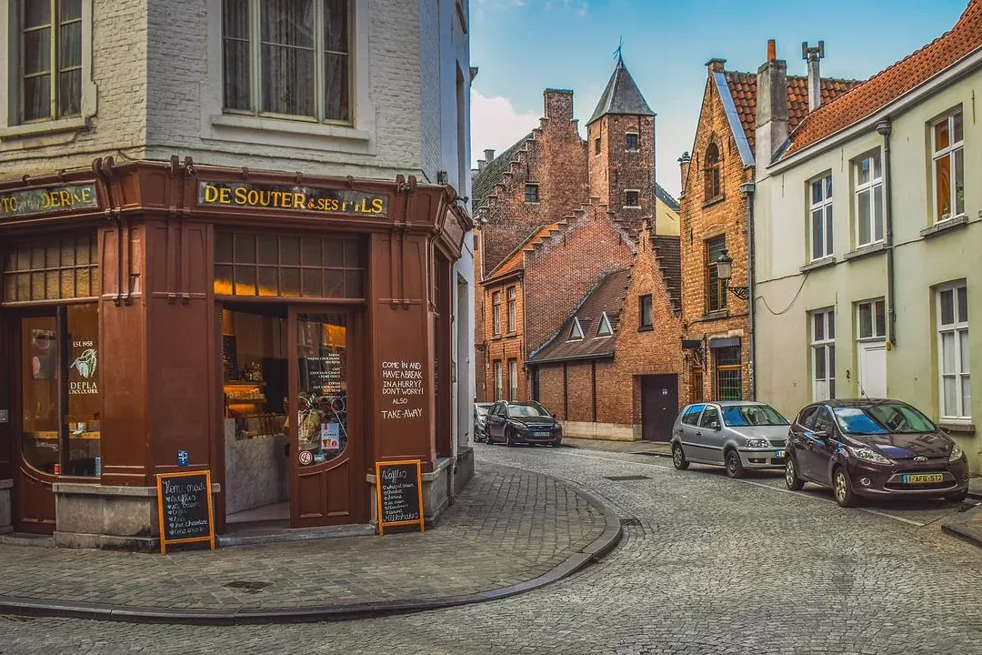 A historic street corner in Bruges with a brown shop on the left, cobblestones, parked cars, and traditional brick buildin...