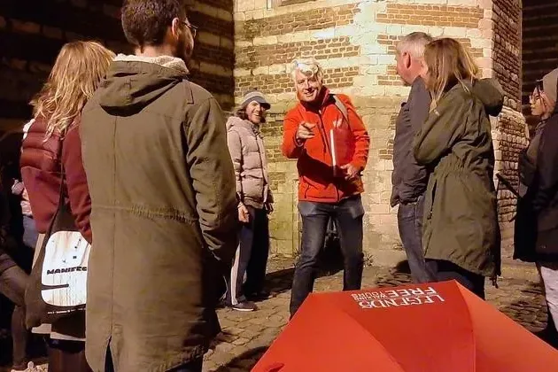 Ein Reiseführer in roter Jacke zeigt auf eine Gruppe von Touristen bei Nacht. Ein roter Regenschirm und eine alte Backstei...