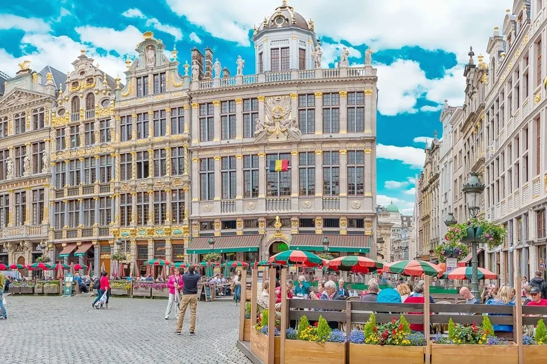 Gremios ornamentados y dorados con bandera belga en la Grand Place de Bruselas, con gente y cafés al aire libre.