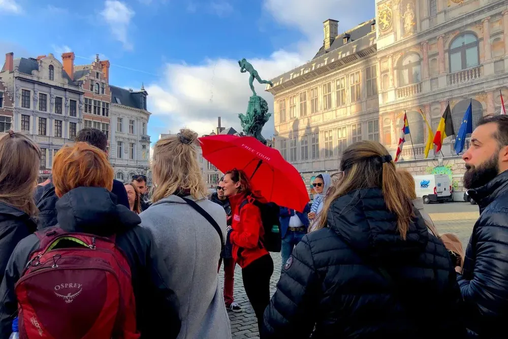 Un grupo de personas escucha a una guía con paraguas rojo en una plaza histórica con la Fuente de Brabo y el Ayuntamiento ...