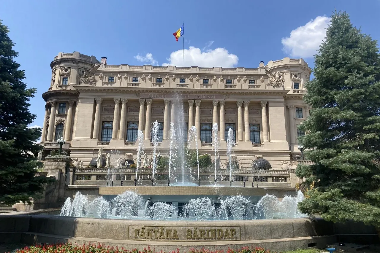 Edificio neoclásico con columnas, torres redondas y bandera rumana, frente a una fuente de varios niveles con chorros de a...