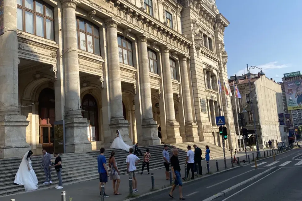 Edificio con columnas, escalones y cúpula. Dos parejas de novios y peatones en acera y escalones. Calle con paso de cebra ...