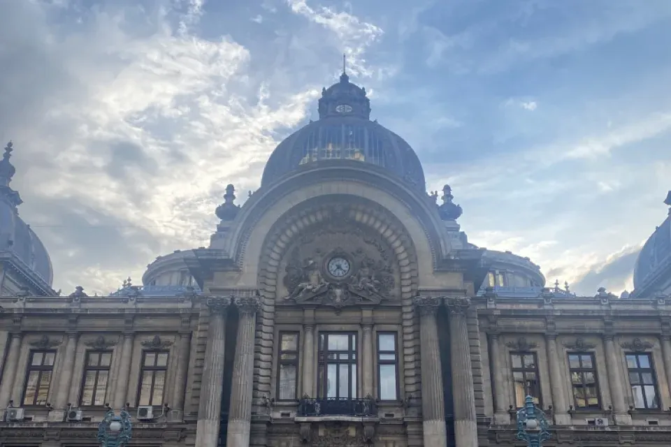 Vista frontal del ornamentado Palacio CEC en Bucarest, un gran edificio con cúpula central, columnas y reloj, con una pers...