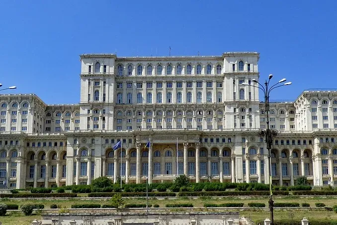 Edificio masivo y ornamentado de piedra blanca con muchas ventanas y columnas bajo un cielo azul claro, visto a distancia ...