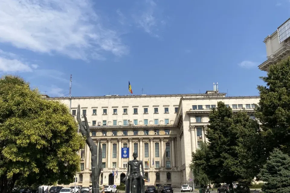 Edificio grande con bandera rumana, dos esculturas modernas oscuras en una plaza pavimentada y árboles verdes bajo un ciel...