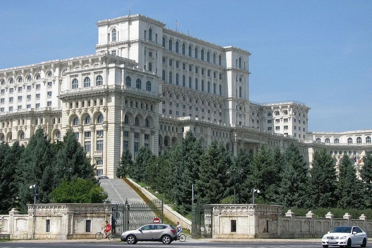 Imponente Palacio del Parlamento en Bucarest, parcialmente cubierto por árboles, con una calle y dos coches.