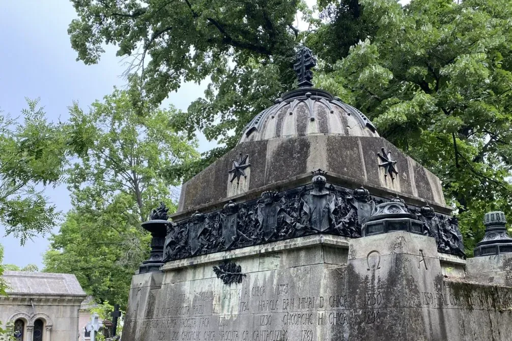 Mausoleo de piedra ornamentado con cúpula, cruz, esculturas en relieve detalladas e inscripciones, rodeado de árboles verd...