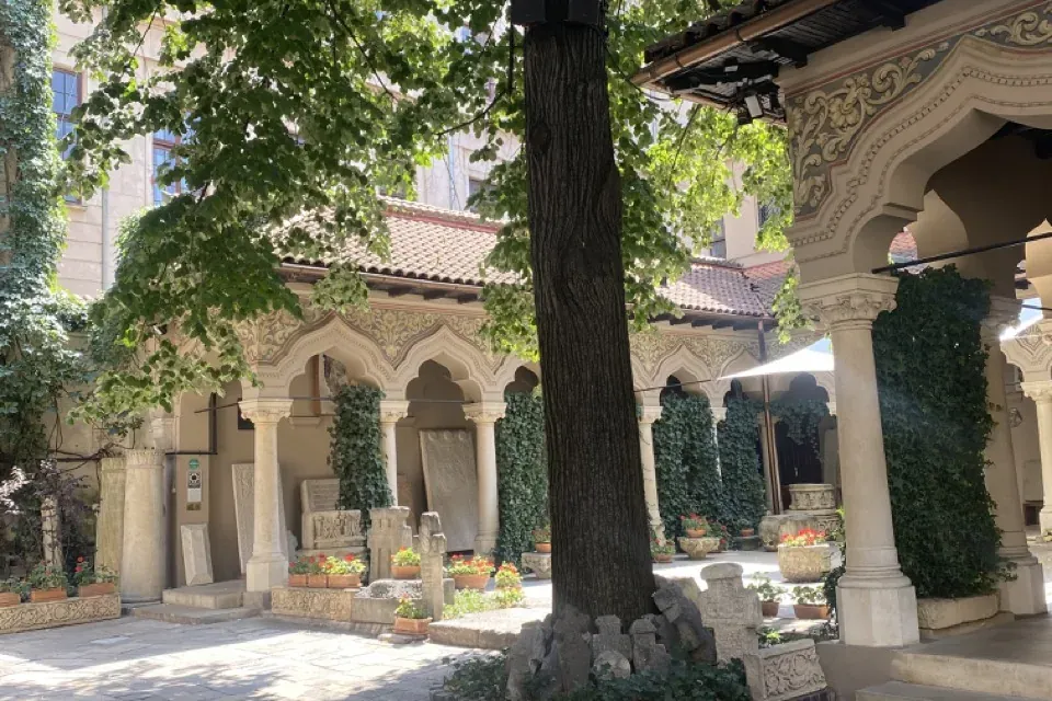 Patio empedrado de un monasterio con un gran árbol, columnatas arqueadas y varias esculturas antiguas de piedra.