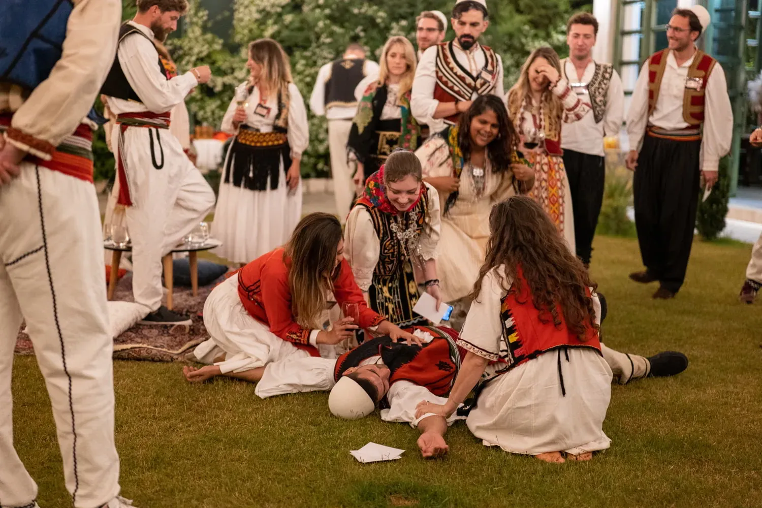 Ludzie w tradycyjnych albańskich strojach uczestniczą w plenerowym występie kulturalnym na trawie. Dwie kobiety klęczą prz...