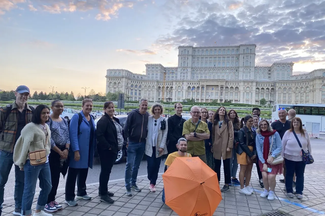 Un grupo diverso de personas de pie frente al enorme Palacio del Parlamento en Bucarest bajo un cielo nublado.