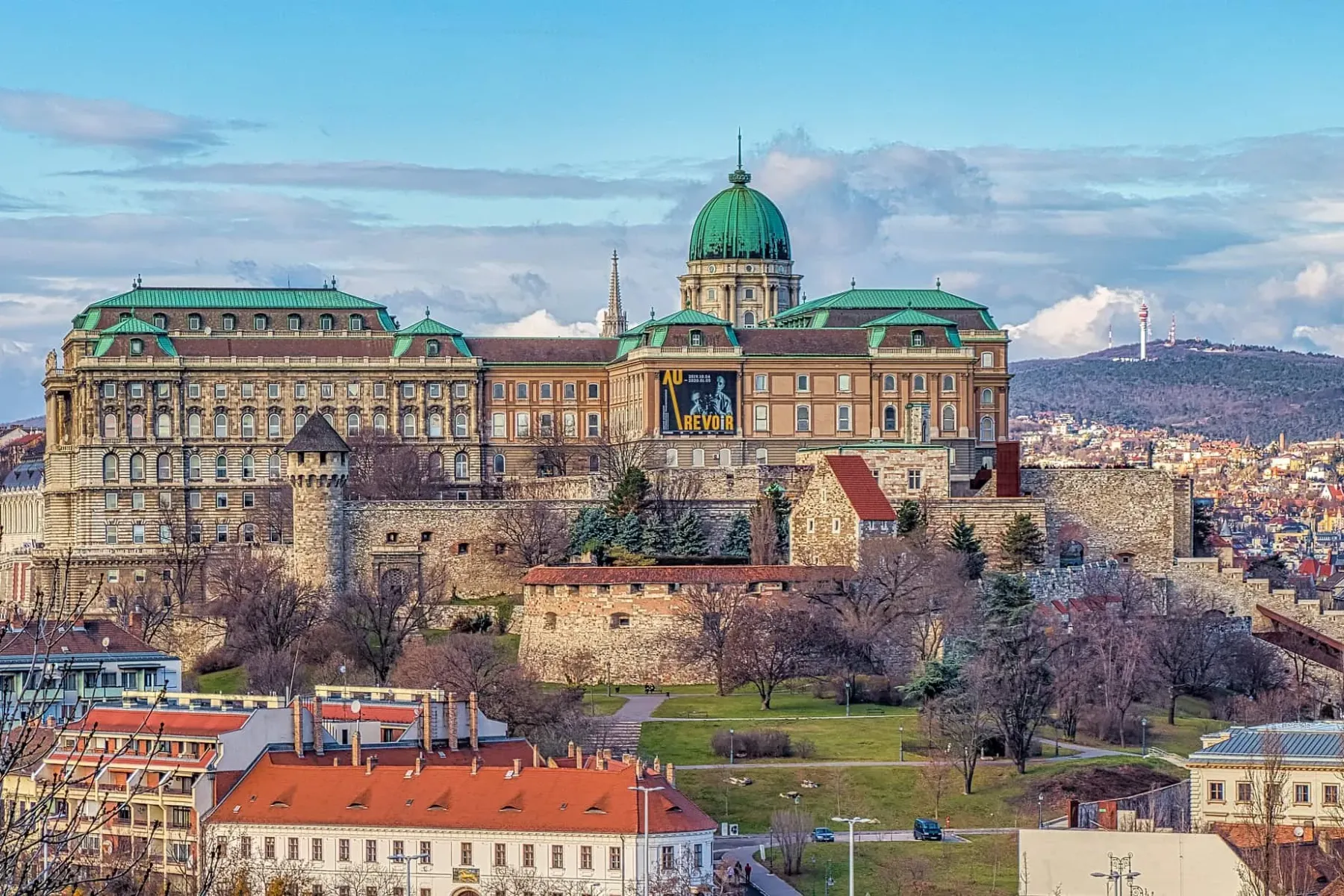 Panoramic view of Buda Castle in Budapest, featuring its grand palace, green dome, stone walls, city buildings, and a dist...