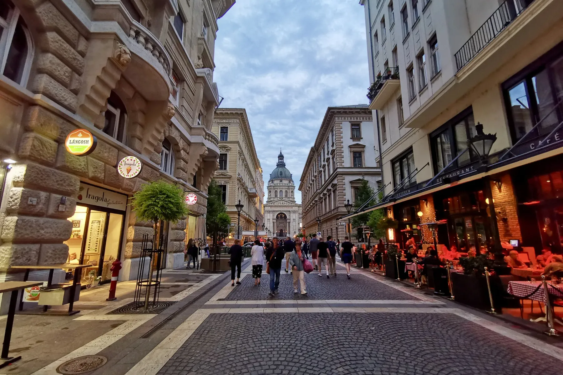 Cobblestone street in Budapest leading to St. Stephen's Basilica, with people walking and outdoor cafes on the sides.