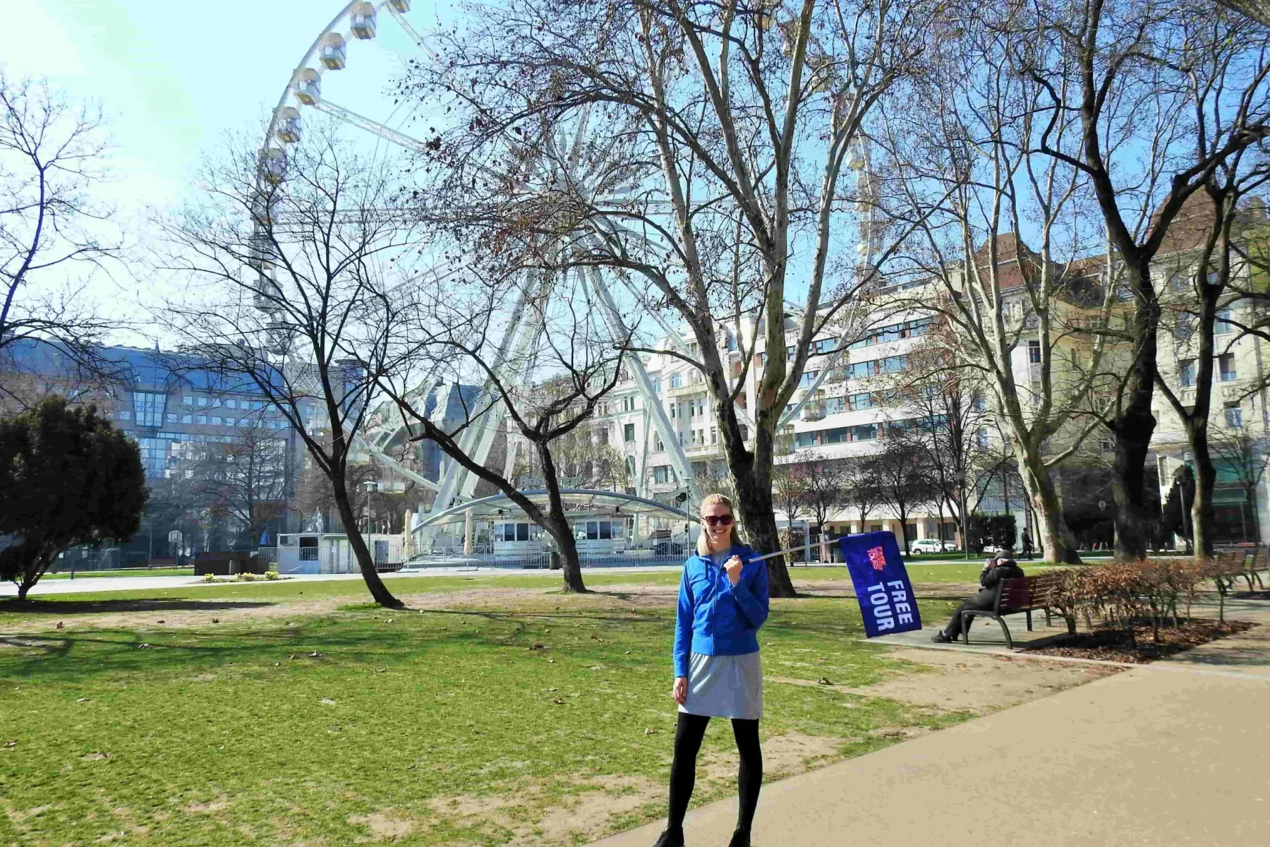 Woman tour guide holding a blue 'Free Tour' flag in a park with a Ferris wheel and leafless trees in Budapest.