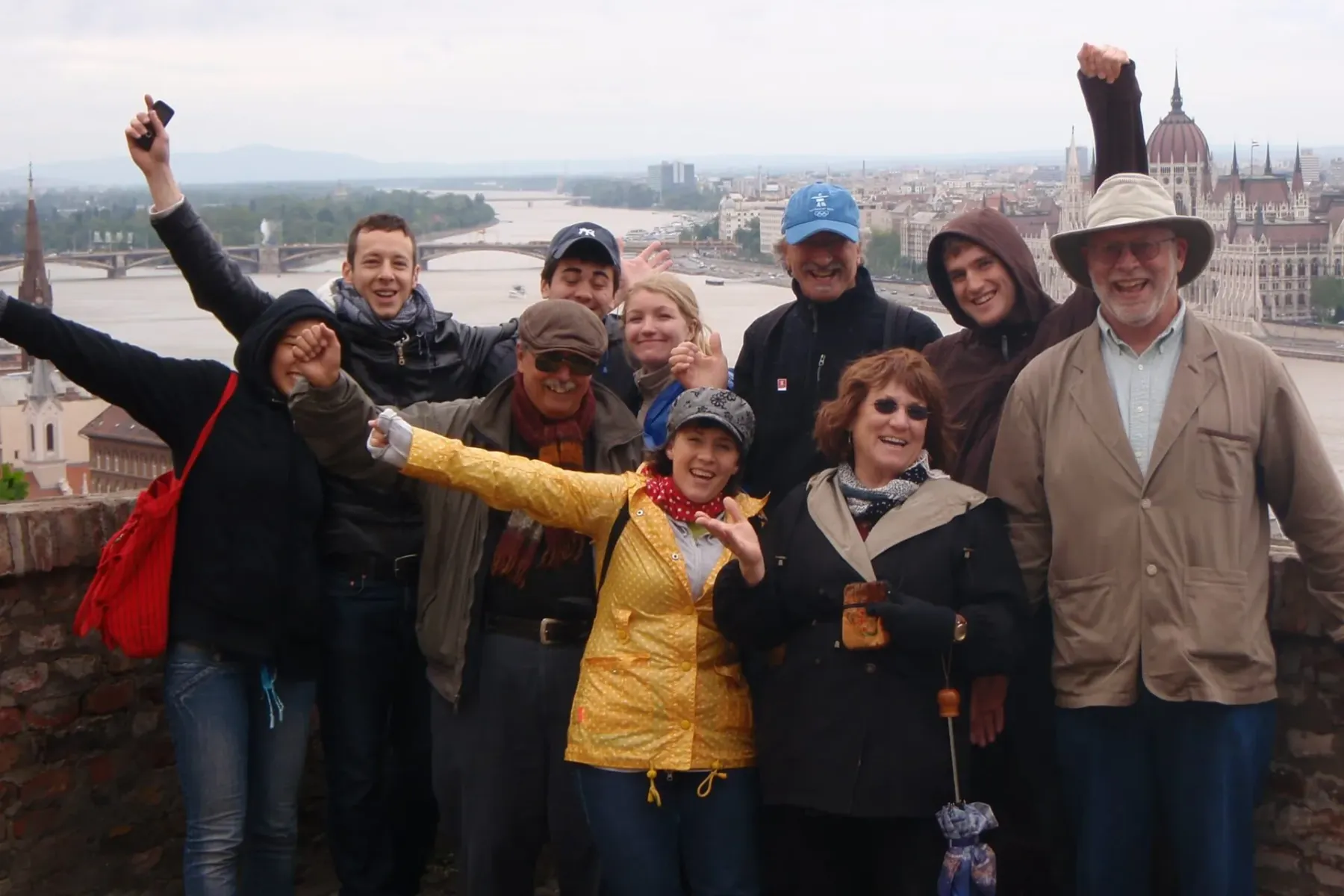 Diverse group of smiling tourists posing on a brick wall with Budapest's Danube River and Parliament building in the backg...