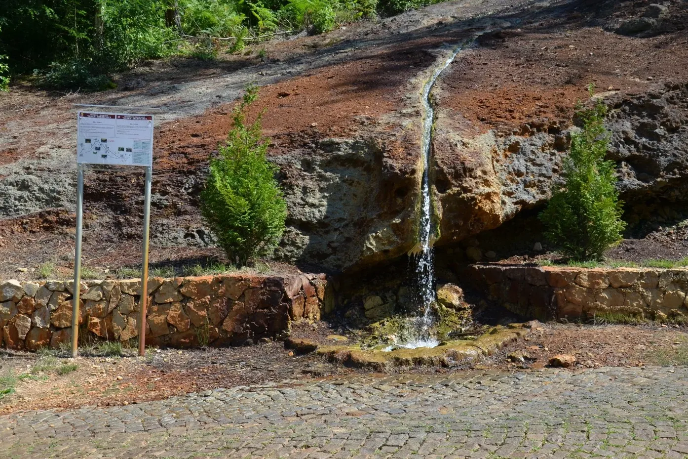 Water flows down a rocky, reddish-brown and grey slope, pooling into a small basin. A stone wall, sign, and small tree are...