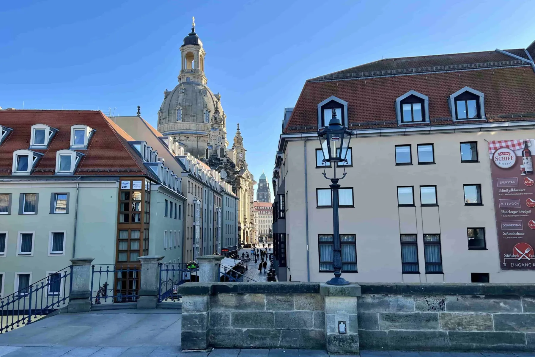 View of Dresden Frauenkirche dome and spire, historic buildings with red roofs, and people on a street, under a clear blue...