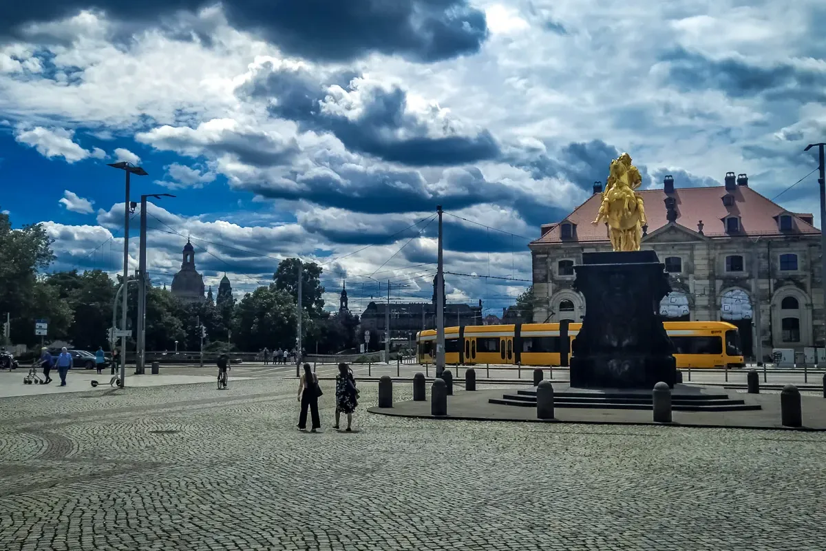 Goldener Reiter Reiterstatue in Dresden auf einem Kopfsteinpflasterplatz, gelbe Straßenbahn, Stadtkuppeln unter bewölktem ...