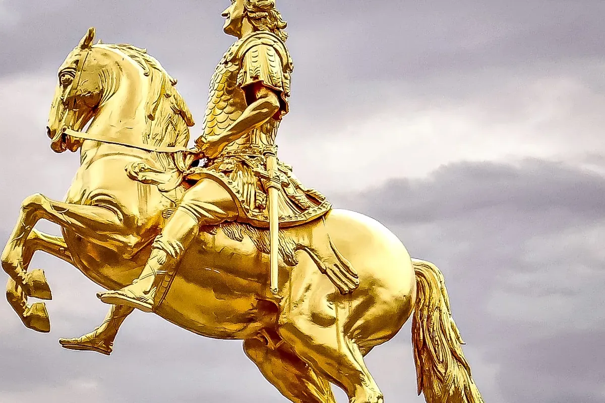 Goldene Reiterstatue eines Mannes in Rüstung auf einem steigenden Pferd vor bewölktem Himmel.