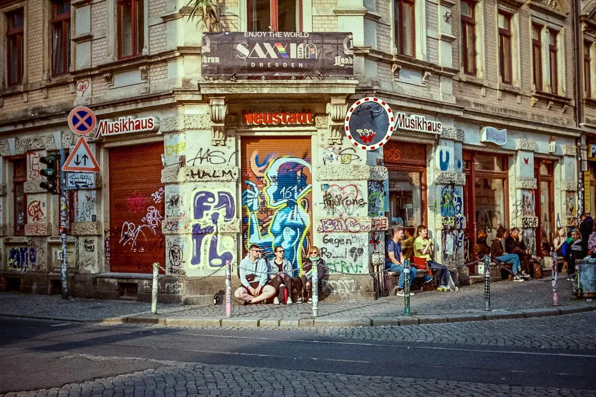 Gebäudeecke in Dresdens Neustadt mit farbenfroher Straßenkunst und Graffiti. Menschen sitzen auf dem Bürgersteig.