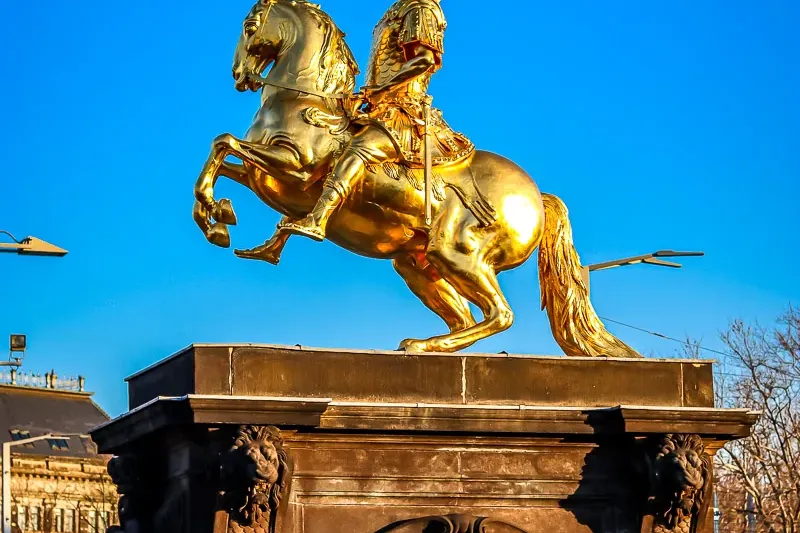 Estatua ecuestre dorada de un hombre a caballo encabritado sobre un pedestal oscuro con inscripciones, bajo cielo azul.