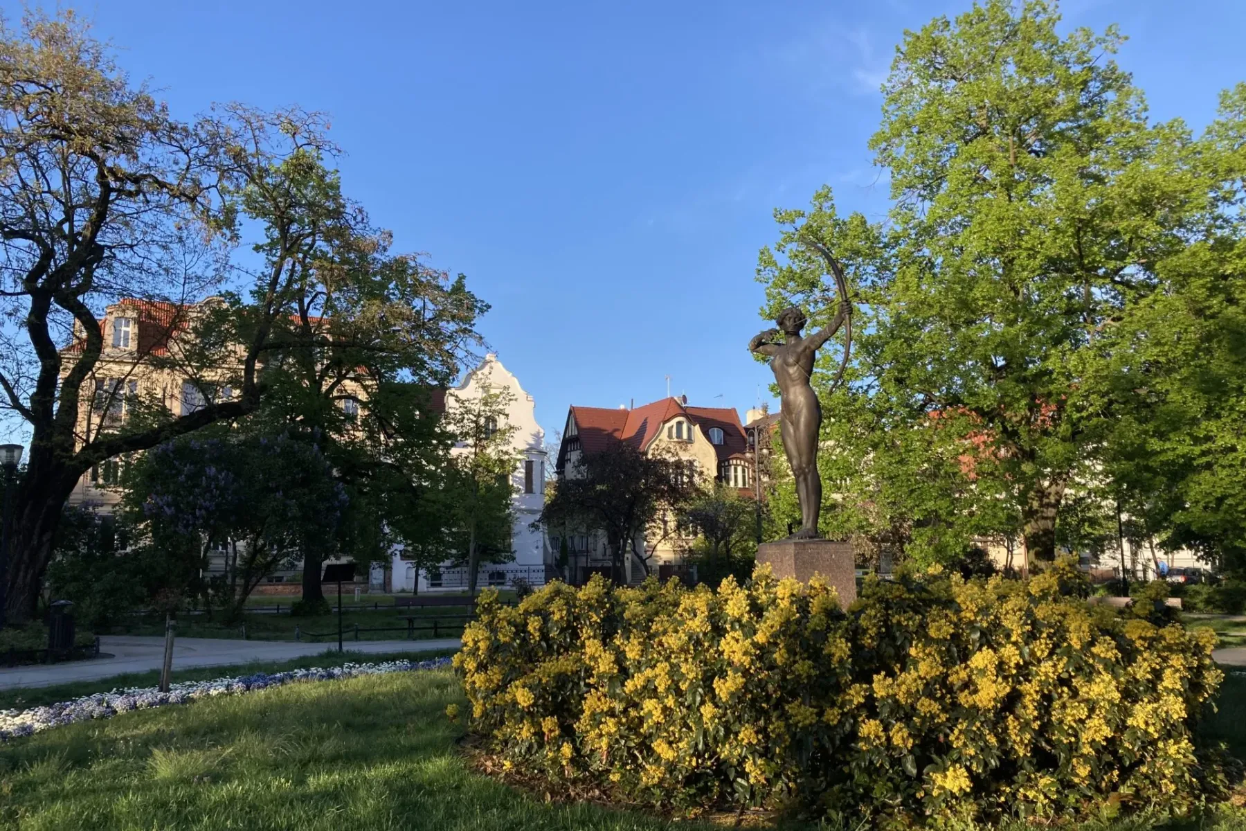 Bronzestatue einer Bogenschützin in einem Park mit gelb blühenden Büschen, grünem Gras, Bäumen und historischen Gebäuden u...