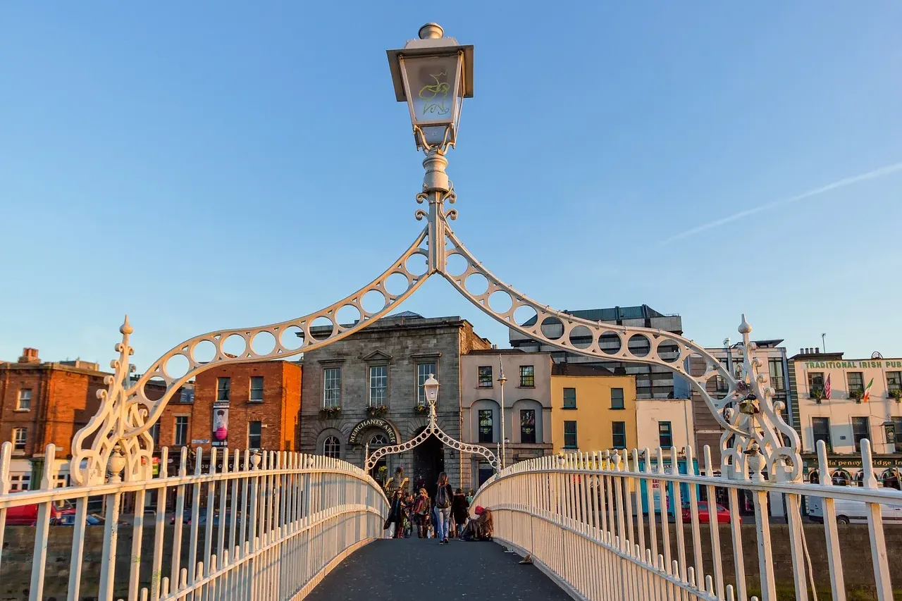 Biały żeliwny Most Ha'penny w Dublinie o zachodzie słońca, z pieszymi i zabytkowymi budynkami nad rzeką Liffey.
