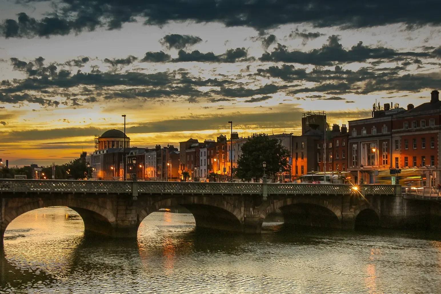Kamienny most łukowy nad rzeką w Dublinie o zachodzie słońca, z oświetlonymi budynkami miejskimi na dalekim brzegu.