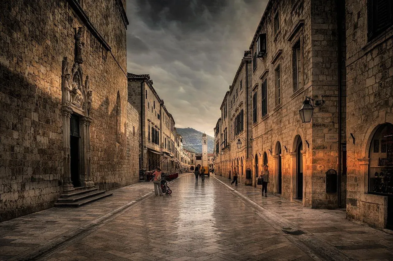 Calle adoquinada mojada en el Casco Antiguo de Dubrovnik, flanqueada por edificios históricos de piedra. Un campanario es ...